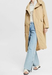 Persona che indossa un trench beige, un top bianco a coste, jeans a vita alta di un leggero blu con spacco frontale e sandali bianchi aperti davanti.