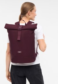 Zaino roll-top color marrone, realizzato in tessuto resistente, con spallacci regolabili, chiusura a gancio e un piccolo logo sulla parte anteriore.