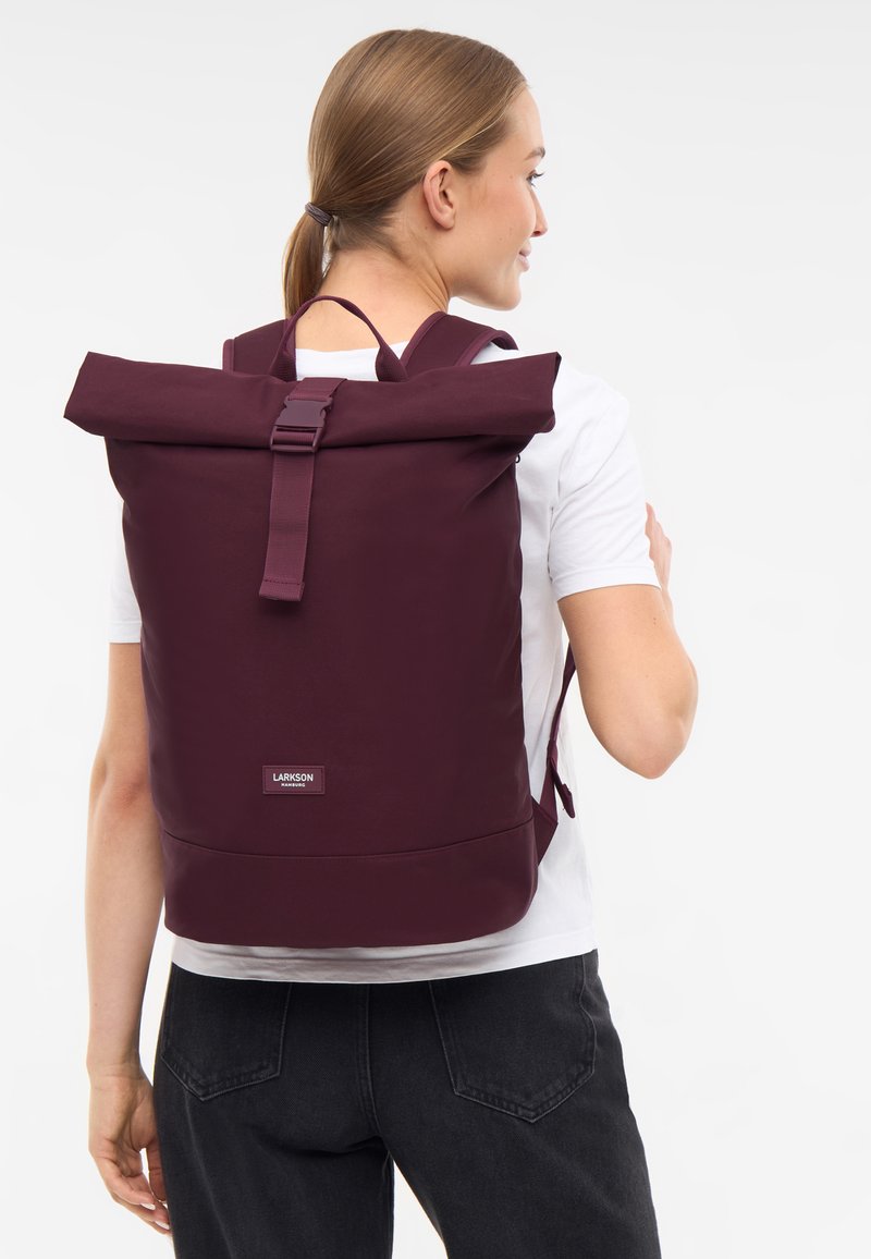 Zaino roll-top color marrone, realizzato in tessuto resistente, con spallacci regolabili, chiusura a gancio e un piccolo logo sulla parte anteriore.