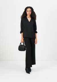 Vrouw met krullend haar staat, draagt een zwarte V-hals blouse, zwarte wijde broek, zwarte veterschoenen met hoge hakken en houdt een kleine zwarte handtas vast.