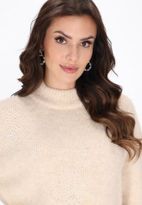 Pulover de culoare crem, cu un guler înalt, având aplicații florale și tricot cu textură. Modelul poartă cercei mari din cercuri clare.