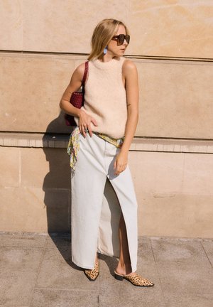 Femme portant un haut beige sans manches en tissu duveteux, une jupe longue blanche fendue sur le côté, des mules imprimé léopard, portant un sac rouge, debout contre un mur de pierre.