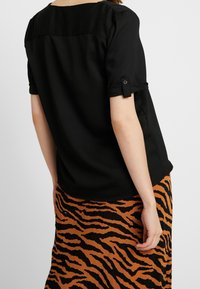 Kvinde iført en sort kortærmet top med knap- og flapærmer og en brun og sort tigerstribet nederdel, set fra ryggen.