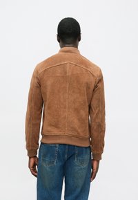 Muž otočený chrbtom, oblečený v hnedej semišovej bundě s rebrovanými manžetami a lemovaním, v kombinácii s modrými riflami na pozadí so svetlým bielym pozadím.