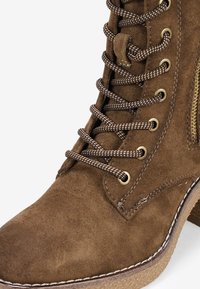 Stivale alla caviglia in suede marrone con superficie texturizzata, occhielli dorati, lacci marroni e tacco basso. Cerniera laterale per un facile accesso e cuciture di dettaglio.