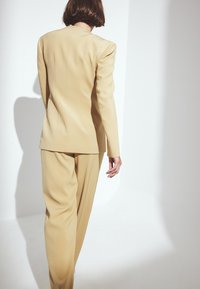 Costume jaune clair avec une coupe ajustée. Comprend un blazer structuré et un pantalon ample, mettant en valeur un tissu lisse et texturé ainsi que des lignes épurées.
