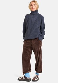 Pull en maille bleu marine avec col montant et fermeture éclair, pantalon en velours côtelé marron, chaussettes bleu clair et chaussures à enfiler multicolores avec un motif de vache.