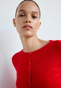 Jeune femme aux cheveux lissés en arrière portant un pull rouge vif boutonné sur un fond blanc uni.