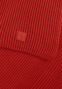 Écharpe en tricot rouge avec une texture côtelée. Présente une petite étiquette carrée avec un motif floral dans un coin. Tissu doux et chaud.