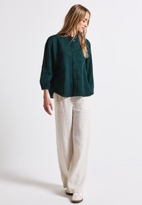 Dark green button-up shirt with long sleeves paired with loose-fitting, light beige trousers and white sneakers. Smooth fabric, relaxed silhouette.