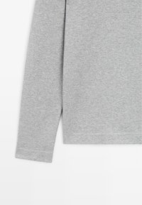 Chemise à manches longues gris clair en tissu doux, montrant les détails des manches et de l'ourlet inférieur sur fond blanc.