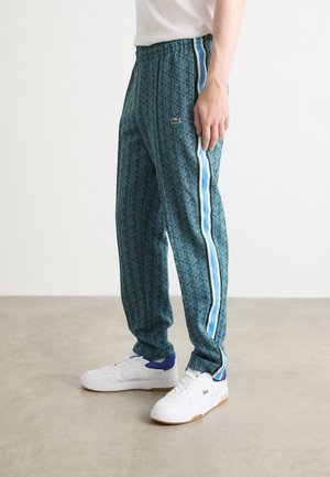 Person trägt grüne, gemusterte Jogginghosen mit blauen und weißen Seitenstreifen und weiße Sneakers mit blauen Akzenten, steht auf einem Betonboden.