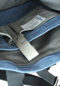 Innenansicht eines Canvas-Rucksacks in Grau und Blau, ausgestattet mit einem Stoffetikett, einem grauen Riemen mit einem Metallschnappverschluss und einem kontrastierenden Reißverschluss entlang der Kante.