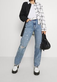 Camicia oversize a scacchi bianco e nero, t-shirt bianca, jeans blu strappati con macchie di vernice e borsa nera. Scarpe platform bianche.