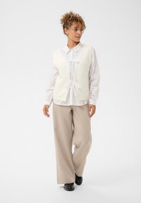 Kobieta o kręconych włosach, ubrana w białą bluzkę, bezrękawnik dzianinowy z kokardkami, beżowe szerokie spodnie i czarne buty, idąca i patrząca na bok.