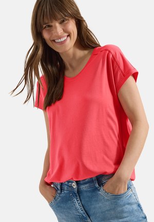 Femme souriante aux cheveux bruns portant un top corail à manches courtes et un jean bleu, les mains dans les poches sur un fond uni.