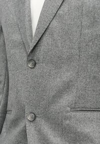 Šedý textura blazer s vykrojeným límcem, dvěma černými knoflíky a bočními kapsami. Tkanina má jemný vzor tkaní.