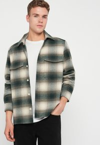 Camisa de franela con un patrón a cuadros en verde, beige y blanco, dos bolsillos en el pecho y cierre de botones. Llevada sobre una camiseta blanca, diseño casual.