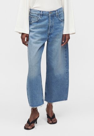 Personne portant un jean bleu clair taille haute à jambes larges avec des ourlets bruts, associé à des sandales à talons noires et un haut blanc à manches longues.