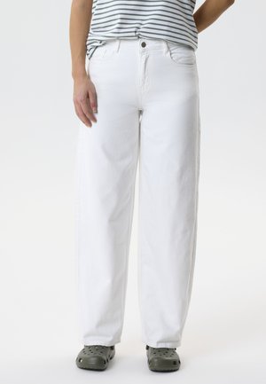 Személy bő, fehér farmert, csíkos inget és sötétszürke papucsot visel, és egyszínű világos háttér előtt áll.