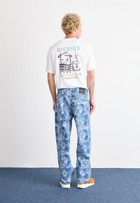Personne avec des cheveux bouclés portant un T-shirt blanc Dickies avec un imprimé industriel, un jean à motifs bleus et des baskets orange, se tenant sur un tapis bleu.