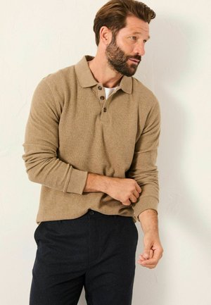 Beige Langarm-Polohemd mit Kragen, einer Knopfleiste mit vier Knöpfen und strukturiertem Stoff. Kombiniert mit dunkelblauen Hosen.