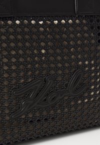 Juodas pintas pirkinių krepšys su lygios odos juosta viršuje ir ryškiu išgaubtu logotipo ženklu. Tekstūrinis raštas suteikia gylio.