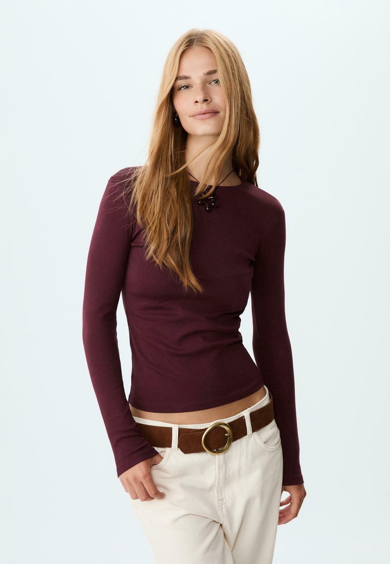 Maroon long-sleeve top with a round neckline, form-fitting design, paired with a brown belt featuring a round gold buckle and light-colored trousers.