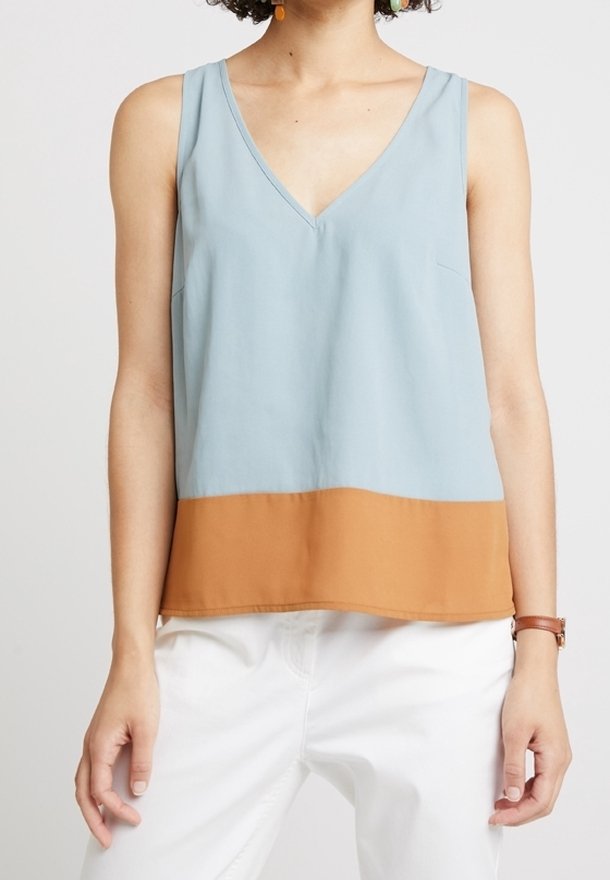Femme portant un haut sans manches à col en V avec une partie supérieure bleu clair et une partie inférieure marron, associée à un pantalon blanc et une montre-bracelet marron.