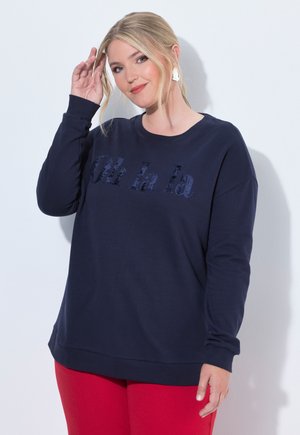 Frau mit blondem Haar trägt einen marineblauen Pullover mit dem Text "Oh la la" und roten Hosen, posiert vor weißem Hintergrund.