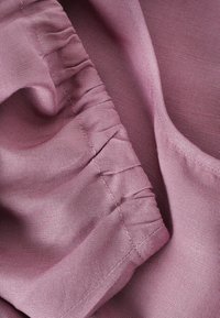 Tessuto rosa chiaro con una superficie liscia e dettagli arricciati. Il materiale è morbido con una leggera lucentezza, che mette in risalto i bordi cuciti.