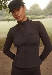 Jachetă sportivă neagră cu guler înalt, închidere cu fermoar frontal și buzunare cu fermoar. Textură netedă, design ajustat și mâneci lungi.