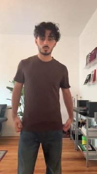 Un giovane con capelli neri ricci indossa una maglietta marrone e jeans blu, in piedi in un soggiorno con piante e librerie.