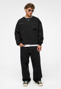 Zwarte sweater met een voorkantzak, gecombineerd met zwarte oversized spijkerbroek en zwarte sneakers met witte accenten. Model met krullend haar en zonnebril.
