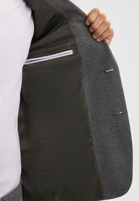 Interno di giacca grigia con una fodera liscia scura, dotato di una tasca con zip e dettagli di cucitura. Tessuto notevole con un esterno a motivo.