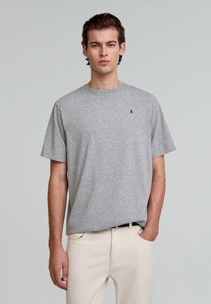 T-shirt en coton gris à manches courtes, col rond, avec un petit logo bleu marine sur la poitrine. Porté avec un pantalon de couleur claire.