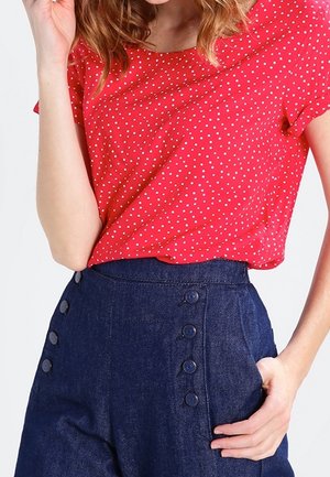 Femme portant un haut rouge à manches courtes avec des pois blancs, rentré dans un pantalon bleu marine taille haute avec deux rangées de boutons.