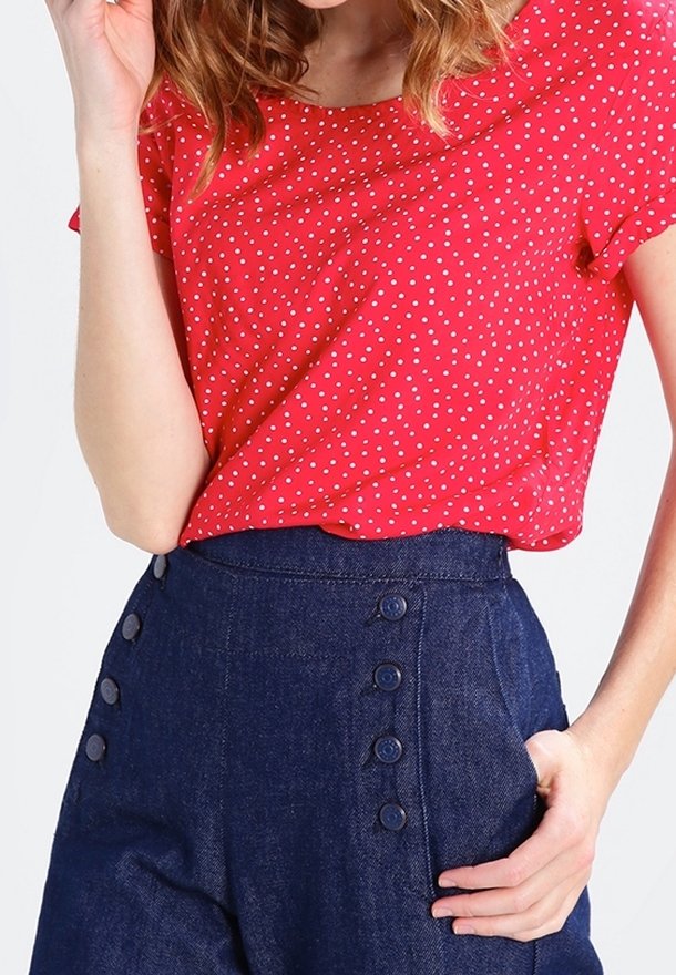 Femme portant un haut rouge à manches courtes avec des pois blancs, rentré dans un pantalon bleu marine taille haute avec deux rangées de boutons.