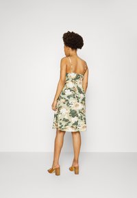 Vestido com padrão floral, alças finas, corpete ajustado e saia rodada. As cores incluem verde, bege e um tom subtil de rosa. Textura de tecido suave.