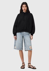 Felpa nera con cappuccio e logo, abbinata a denim shorts azzurri chiari con dettagli strappati e sandali neri con lacci.
