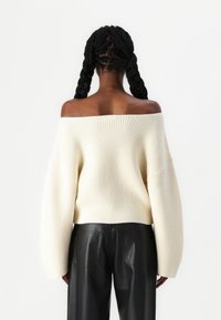 Sweater off-white à épaules dénudées avec texture côtelée, manches larges et coupe ample, associé à un pantalon en cuir noir taille haute.