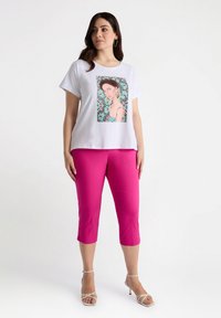 Hvid t-shirt med korte ærmer og farverigt grafiktryk, parret med lyserøde cropped bukser og sandaler med remme, sat mod en neutral baggrund.