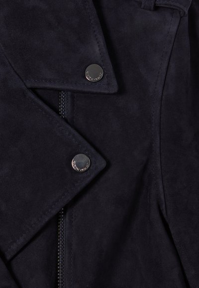 Gros plan sur une veste en daim bleu marine foncé avec des boutons-pression argentés et une fermeture éclair noire visible sous les revers qui se chevauchent.