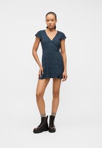 Γυναίκα με κοτσίδες cornrow, που φοράει φορέμα navy blue με λουλούδια και μαύρα δερμάτινα αρβυλάκια με κορδόνια, στέκεται μπροστά σε λευκό φόντο.