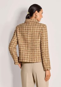 Vrouw met donker haar in een lage paardenstaart, gekleed in gouden oorbellen, een gestructureerde bruine tweedjas en beige hoge taillebroek, met de rug naar de kijker gekeerd.