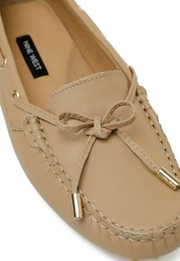 Beigeleder-Mokassin-Schuh mit einer Schleifendetails, goldfarbenen Hardwareakzenten und kontrastierenden Nähten entlang der Kanten. Weiche Textur.