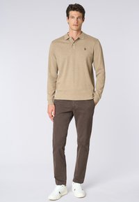 Polo a maniche lunghe beige con colletto, caratterizzato da un piccolo logo. Abbinato a pantaloni marroni scuri e sneaker bianche, su uno sfondo semplice.