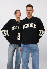 Black knit sweatshirts featuring cream "ICONIC" text, number "33" on sleeves, and distressed hems, paired with light blue jeans.
