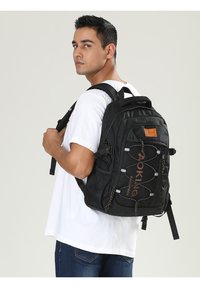 Zaino nero con spallacci regolabili, cordini elasticizzati sul davanti e tessuto testurizzato. Include un grande patch con logo e diversi scomparti.