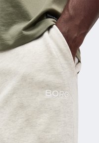 Άτομο με σκούρο χρώμα δέρματος με το χέρι στην τσέπη ανοιχτό μπεζ φόρμας, φοράει ελαφρώς πράσινο πουκάμισο με ορατή ετικέτα "BJÖRN BORG" και κέντημα "BORG".